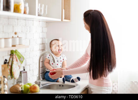 Adorable baby sitting on table in kitchen while mom washing apple Stock Photo
