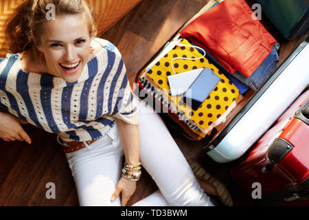 Upper view of smiling elegant woman in white pants and striped blouse sitting near open travel suitcase in the modern living room in sunny summer day. Stock Photo