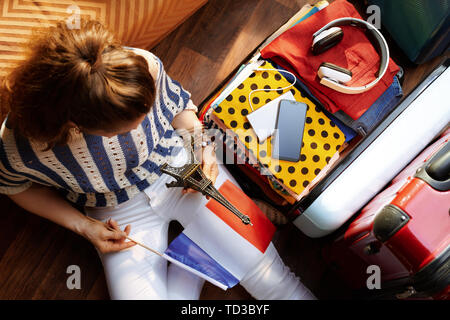 Upper view of stylish woman in white pants and striped blouse with French flag and Eiffel tower souvenir near open travel suitcase at modern home in s Stock Photo