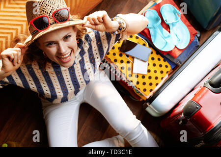 Upper view of smiling elegant woman in white pants and striped blouse with supper hat and sunglasses near open travel suitcase in the modern living ro Stock Photo