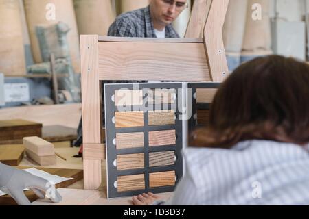 Furniture production master making wooden chair, female designer with wood samples choosing finishing in woodworking workshop. Stock Photo