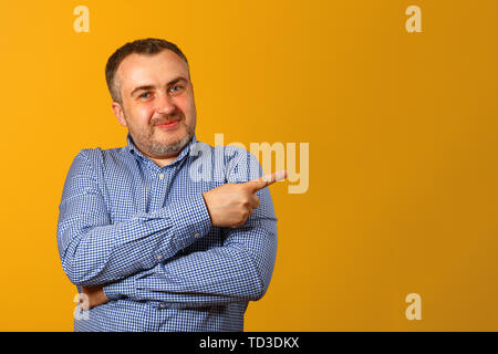 Handsome bearded man in a shirt, points his finger to the side, looks into the camera and smiles, against a yellow background. Copy space. Close-up. Stock Photo
