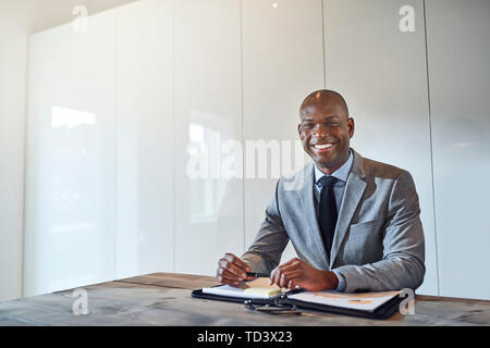Smiling young African American businessman sitting alone at a table in a boardroom with a folder full of paperwork Stock Photo