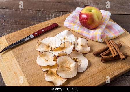 Front view Dried apple chips , raw apple , cinnamon sticks on cooking board over wood background. Stock Photo