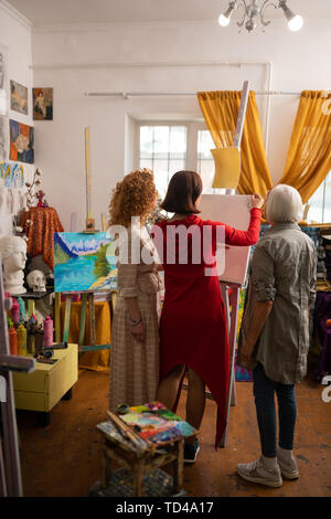 Looking at students. Grey-haired aged artist looking at her talented students drawing on canvas Stock Photo
