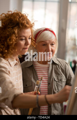 Student feeling involved. Curly red-haired student feeling involved in painting standing near tutor Stock Photo