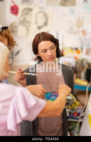 Standing near teacher. Dark-haired art student holding painting brush standing near teacher Stock Photo