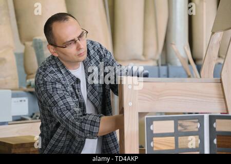 Male carpenter working in furniture joinery, master making wooden chair in wood workshop. Stock Photo