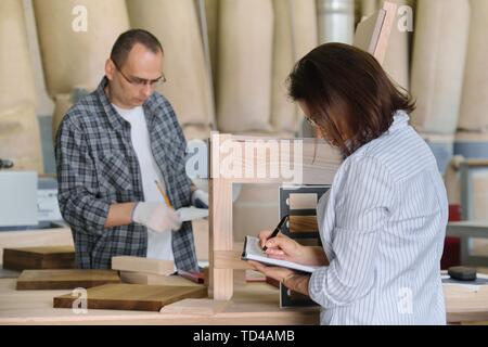 Furniture production master making wooden chair, female designer with wood samples choosing finishing in woodworking workshop. Stock Photo