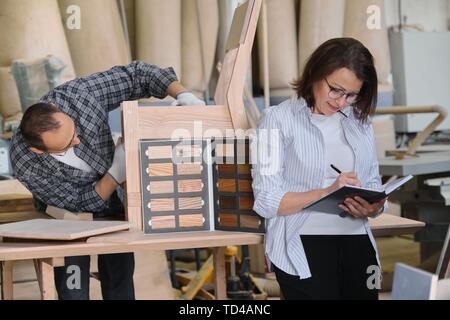 Furniture joinery production, working male joiner and female designer with wood samples choosing finishing, writing in business notebook. Stock Photo