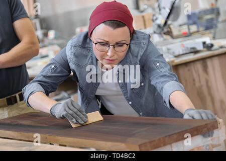 Mature craftswoman working in woodshop. Female painting wooden board with oil, varnish. Furniture joinery wood business. Stock Photo