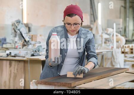 Middle-aged craftswoman working in woodworking workshop. Female varnishing wooden board with oil, varnish. Furniture joinery wood business. Stock Photo