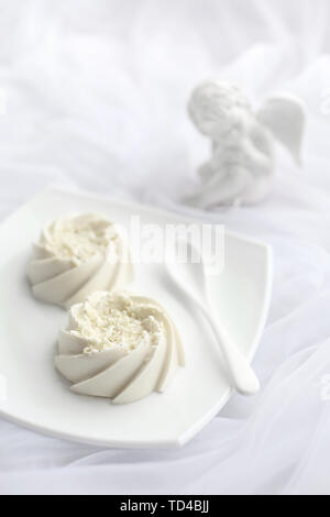 Light airy dessert panna cotta mousse with white chocolate and vanilla on top, on white ceramic plate and ceramic spoon and porcelain angel statuette. Monochrome Stock Photo