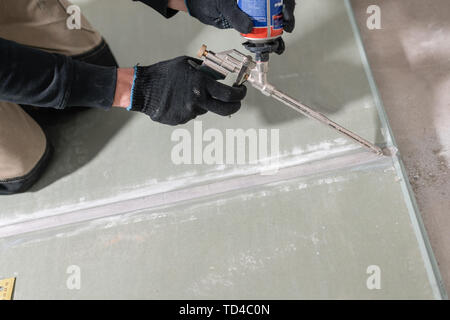 Worker is using a polyurethane foam for gluing drywall at ninety degrees. Hand holding polyurethane expanding foam glue gun applicator Stock Photo