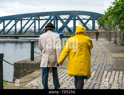 The Shore, Leith, Edinburgh, Scotland, United Kingdom, 12th June 2019. UK Weather: A couple holding hands walking on the riverbank on a cold rainy and windy day along the Water of Leith river Stock Photo