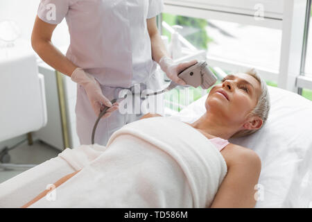 Professional cosmetology. Serious aged woman lying on the medical bed while having anti age facial treatment procedure Stock Photo