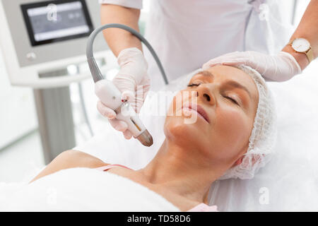 During the procedure. Nice mature woman lying with her eyes closed while having a neck lifting procedure Stock Photo