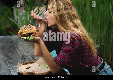 Student consumes fast food. Girl bites a very big burger in a garden. Stock Photo