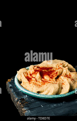 a homemade hummus seasoned with paprika served in a green ceramic plate, on a dark gray rustic wooden table against a black background with some blank Stock Photo