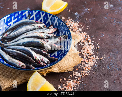 Blue fish. Fresh anchovies in a dish with pink salt and lemon on dark background. Healthy protein meal. Local seafood, Italy Stock Photo