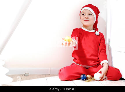 A little boy in the shape of Santa Claus is smiling, stands on a white background, and holds a tangerine in his hand Stock Photo