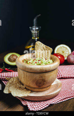 delicious guacamole in a wood bowl with tortilla chips next to avocado and other ingredients, typical mexican healthy vegan cuisine with rustic dark f Stock Photo