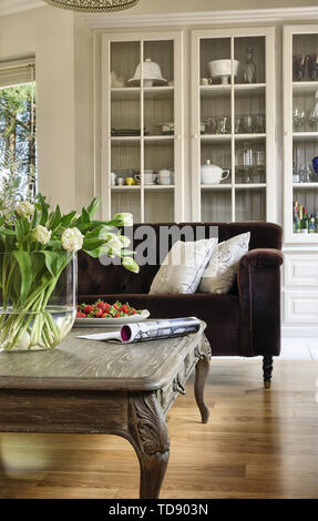 White tulips in large clear glass vase on wooden coffee table in front of tufted sofa in living room    UK AND IRISH RIGHTS ONLY Stock Photo