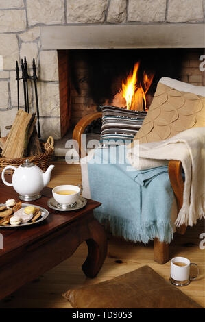 Teapot and biscuits on coffee table with chair in front of fireplace with lit fire    UK AND IRISH RIGHTS ONLY Stock Photo