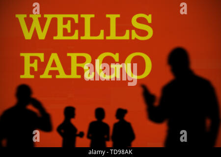 The Wells Fargo logo is seen on an LED screen in the background while a silhouetted person uses a smartphone in the foreground (Editorial use only) Stock Photo