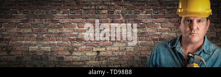 Male Contractor In Hard Hat Holding Hammer In Front Of Old Brick Wall Banner with Copy Space. Stock Photo