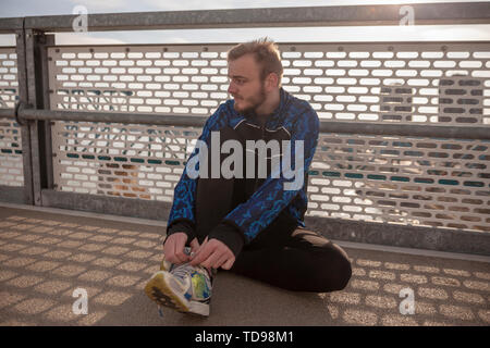 one young man, tying his shoelaces on a running shoes, looking sideways. sitting on a ground (asphalt), leaning to a fence on a bridge. Stock Photo