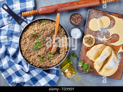 cooking pierogies with ground liver with onion filling fried in a skillet, raw dough and uncooked dumpling on a cutting bord with rolling pin and spic Stock Photo
