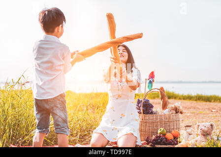 Mother and son play fencing with bread together when picnic at outdoors near lake or river. People lifestyles and Family. Motherhood and childhood con Stock Photo