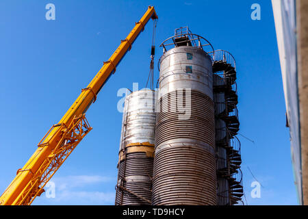 Crane is carry and keeps balance with heavy metal silo in industrial complex. Stock Photo