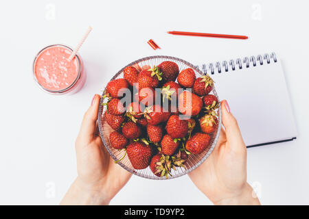 Woman's hands holding bowl of fresh strawberries above white table with empty notebook and smoothie, flat lay. Stock Photo