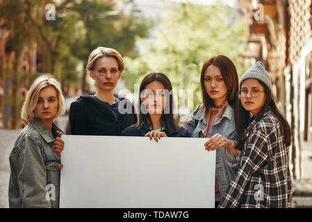 Group of protesting young female activists holding blank signboard and looking at camera while standing on the road. Women's march. Human rights. Prot Stock Photo