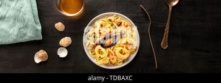 Seafood pasta panorama. Tagliolini with mussels, shrimps, clams and squids, with a glass of white wine, shot from the top on a black background Stock Photo