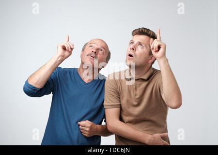 Two men father and son looking upwards, pointing index fingers up with shock and fear . Stock Photo