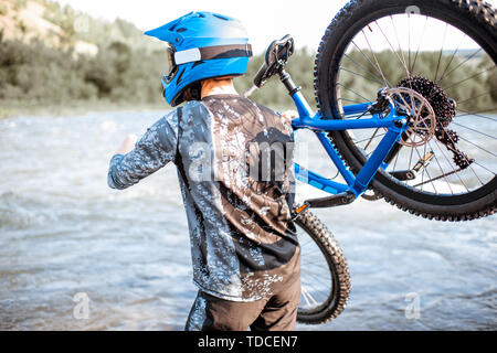 Professional well-equipped cyclist standing with bicycle on the riverside in the mountains. Concept of a freeride and off road cycling Stock Photo