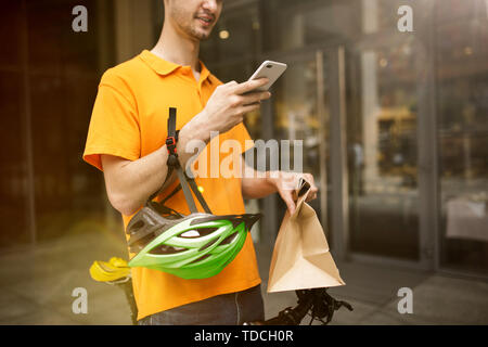 Young man in yellow shirt delivering package using gadgets to track order at the city's street. Courier using online app for receiving payment and tracking shipping address. Modern technologies. Stock Photo