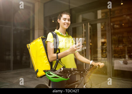 Young woman in yellow shirt delivering food using gadgets to track order at the city's street. Courier using online app for receiving payment and tracking shipping address. Modern technologies. Stock Photo