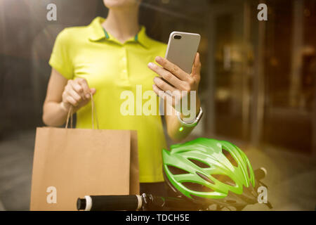 Young woman in yellow shirt delivering package using gadgets to track order at the city's street. Courier using online app for receiving payment and tracking shipping address. Modern technologies. Stock Photo