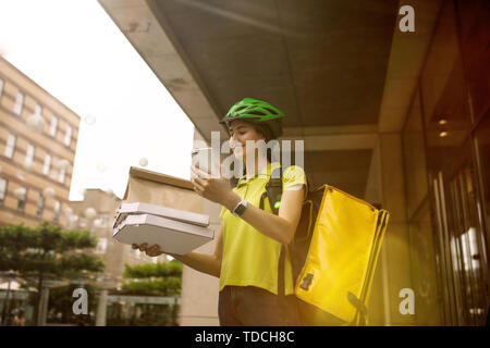 Young woman in yellow shirt delivering pizza using gadgets to track order at the city's street. Courier using online app for receiving payment and tracking shipping address. Modern technologies. Stock Photo