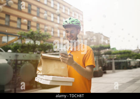 Young man in yellow shirt delivering pizza using gadgets to track order at the city's street. Courier using online app for receiving payment and tracking shipping address. Modern technologies. Stock Photo