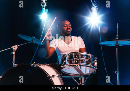 Young african-american jazz musician or drummer playing drums on blue studio background in glowing smoke around him. Concept of music, hobby, inspirness. Colorful portrait of joyful attractive artist. Stock Photo