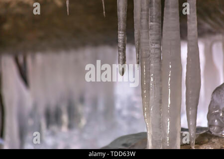 Close up view of smooth and shiny icicles under a bridge in winter. Rocks covered with frozen water can be seen beneath the icicles. Stock Photo