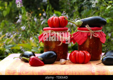 Eggplants in tomatoes in jars are located on a table in the garden, Fresh tomatoes, aubergines, garlic and bell peppers are on the table, blanks for t Stock Photo