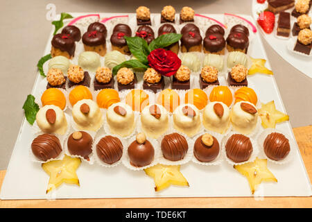 Small assorted cakes lined up decorated on dessert buffet with red rose and mint leaves, with slices of Carambola fruit Stock Photo