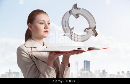 Woman looks at transparent recycle 3d symbol above opened notebook. Renewable energy source concept. Elegant girl with book on background of cityscape Stock Photo