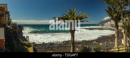 Steep high lava rock cliffs. Turquoise ocean rests on a bright blue sky and a line of clouds above the horizon. Playa Arena beach, Tenerife. Wide angl Stock Photo